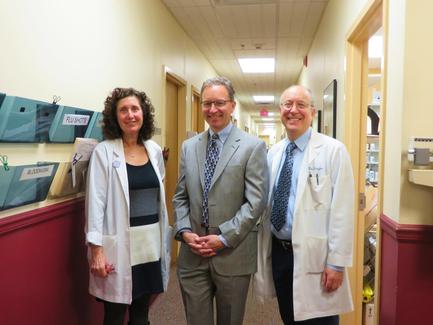 Three people standing in a hallway. On the left is a woman with curly hair wearing a white lab coat. She is standing next to a man in a gray suit and tie. The man in the middle is also wearing a suit and glasses. All three people are smiling and appear to be posing for the photo. On the right side of the image there is a man wearing a blue shirt and tie who is also smiling. He is standing in front of a red wall with a shelf on the left side. The shelf has various medical supplies hanging on it. The hallway appears to be well-lit with natural light coming in from the windows.