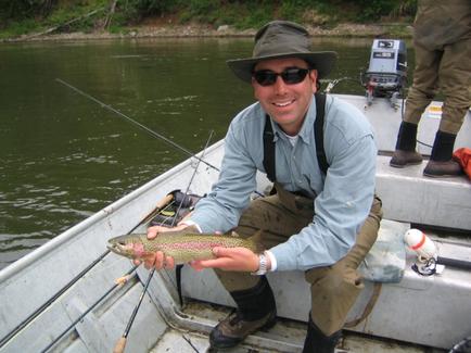 A man sitting in a boat on a river. He is wearing a green hat sunglasses and a blue shirt. He has a big smile on his face and is holding a fish in his hands. The fish appears to be a rainbow trout with a pink and green speckled pattern on its body. There are fishing rods and other equipment on the boat and another person can be seen in the background. The river is calm and there are trees and bushes along the banks.