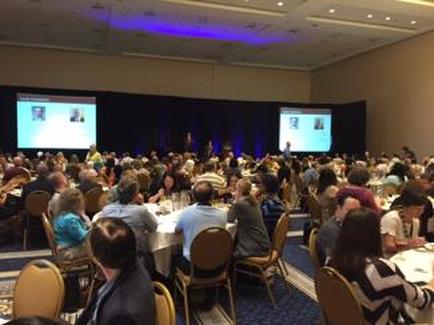 A large conference room with a large number of people sitting at tables facing a stage. The room has a high ceiling with recessed lighting and a carpeted floor. On the stage there are two large screens displaying images and text. The people in the room appear to be engaged in a discussion or presentation. They are all dressed in casual clothing and are seated in rows of chairs facing the stage.