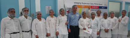 A group of people standing in a room with blue walls and a whiteboard in the background. There are nine people in the group all wearing white lab coats and head caps. They are all smiling and appear to be posing for a photo. In the center of the group is a man wearing a blue shirt and black pants who is standing in front of a sign that reads "Do Check Act". He is holding a clipboard and appears to be giving a presentation or presentation to the group. The people around him are also smiling and looking at the camera.