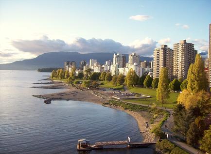An aerial view of a city with a large body of water in the foreground. The water is calm and there is a small pier extending into the water. On the right side of the image there are tall buildings with multiple floors and balconies. The buildings are surrounded by trees and greenery and there are a few people walking along the shore. In the background there is mountains and a clear blue sky. The overall scene is peaceful and serene.