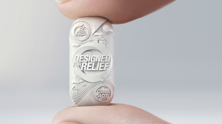 A close-up of a person's finger holding a small round object. The object appears to be made of a light-colored material and has a textured surface. The text on the object reads "Designed for Relief" in a bold sans-serif font. Below the text there is an illustration of a cityscape with buildings trees and a sun. The background is blurred making the object the focal point of the image.