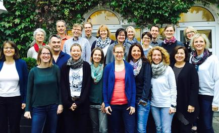 A group photo of a large group of people standing in front of a building with a green wall and arched windows. There are around 20 people in the photo all of them are smiling and looking at the camera. They appear to be of different ages genders and ethnicity's. Some people are wearing scarves while others are wearing casual clothing. The building in the background has a white door and a window with a yellow frame. The overall mood of the photo is happy and relaxed.