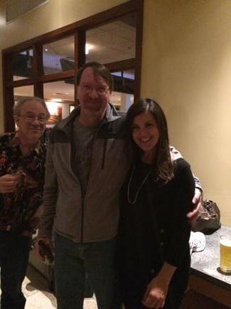 Three people standing in a room with a wooden floor and a window in the background. The person on the left is a man with glasses and a beard wearing a gray jacket and a floral shirt. He is standing next to a woman with long brown hair who is wearing a black jacket and jeans. They are all smiling and appear to be posing for the photo. On the right side of the image there is another person who appears to be an older man holding a glass of beer.