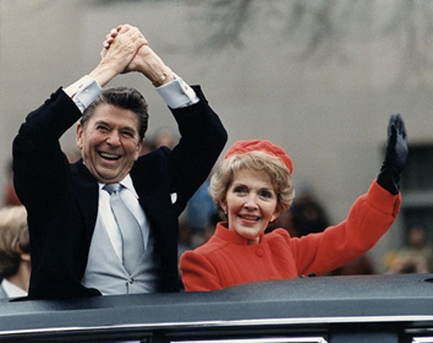 A man and a woman sitting in the back of a car holding hands and smiling. The man is on the left side of the image wearing a black suit and tie and the woman on the right side is wearing a red coat and a red hat. They appear to be in a parade or event as there are other people visible in the background. The car appears to be a vintage model possibly from the early 20th century.