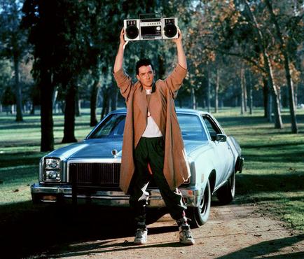 A young man standing in front of a vintage car in a park. He is holding up a boombox above his head with both hands as if he is dancing or celebrating. The man is wearing a long brown coat a white t-shirt and green pants. He has a serious expression on his face and is looking directly at the camera. The car is a silver sedan with a black grille and headlights. The park is filled with trees and there is a grassy lawn in the background. The sky is blue and the overall mood of the image is joyful and carefree.