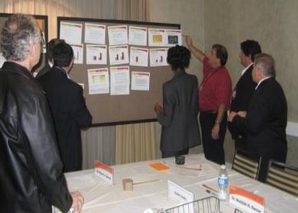 A group of people gathered around a table in a conference room. The table is covered with a white tablecloth and there are nameplates and water bottles on it. On the wall behind the table there is a large bulletin board with multiple papers pinned to it. The papers appear to be related to a presentation or presentation. The people in the image are of different ages and genders and they are engaged in conversation. One person is pointing to one of the papers on the board while another is looking at it intently. The room appears to be well-lit with natural light coming in from the windows.