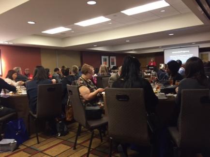 A large conference room with a group of people sitting at tables. The room has a high ceiling with recessed lighting and a projector screen in the background. The people are of different ages and genders and they appear to be engaged in a discussion or presentation. The tables are covered with white tablecloths and there are plates cups and saucers on the tables. There is a woman standing at the front of the room giving a presentation to the audience. The walls are painted in a light color and there is a red carpet on the floor.