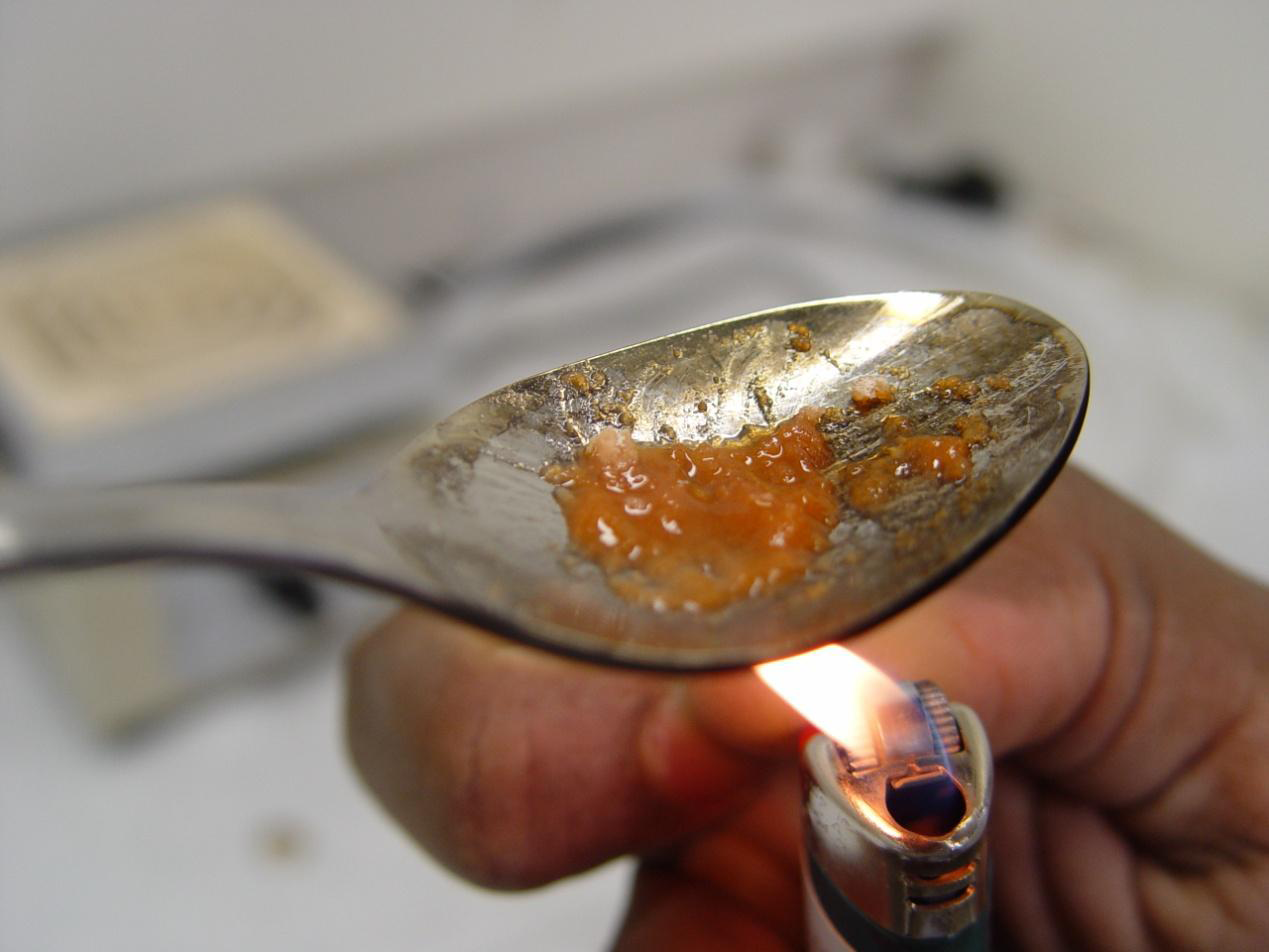 A silver spoon with a small amount of orange-colored substance on it likely an opioid or another drug. A person's hand is holding a lit lighter under the spoon. The background is blurred but it seems to be a kitchen countertop.