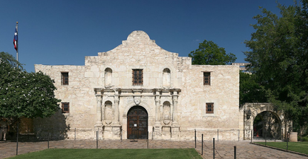 A large stone building with a dome on top. The building appears to be old and weathered with a rough uneven surface. It has multiple arched windows and a large wooden door in the center. The entrance of the building is flanked by two smaller arches on either side. There is a flagpole with the American flag flying in the background. The sky is blue and there are trees and shrubs surrounding the building. The ground is covered in green grass.