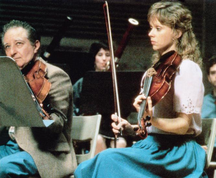 A man and a woman playing violins in an orchestra. The man is on the left side of the image playing the violin with a serious expression on his face. He is wearing a suit and tie and appears to be in the middle of a performance. The woman is sitting next to him holding the violin and looking at him attentively. She has blonde hair and is wearing blue skirt and a white blouse. In the background there are other musicians playing various instruments including a trombone and a cello. The orchestra is seated in rows of chairs and there are music stands in front of them. The image appears to have been taken in a dimly lit room.