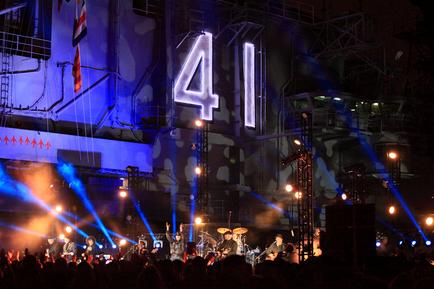 A large stage with a band performing on it. The stage is set up with a large number "41" in the center which is lit up with blue and purple lights. The band members are playing various instruments including guitars drums and keyboards. The audience is visible at the bottom of the image with their hands raised in the air cheering and enjoying the performance. In the background there is a large screen with the number 41 on it and a flag hanging from the ceiling. The overall atmosphere of the stage is energetic and lively.