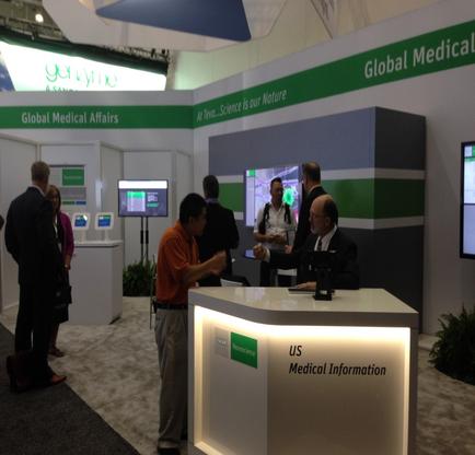 A trade show booth with a green and white color scheme. The booth has a sign that reads "Global Medical Affairs" and "US Medical Information". There are several people standing around the booth some of them are engaged in conversation. In the center of the image there is a man sitting at a desk with a computer monitor in front of him. He is wearing a suit and appears to be explaining something to another man who is standing next to him. There are two large screens on either side of the desk displaying information about the booth. The background shows other booths and people walking around.