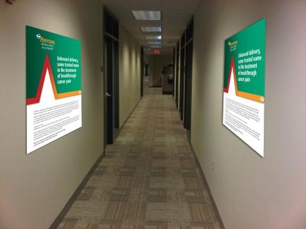 A long corridor with two posters hanging on the wall on either side. The posters have a green background with white text and a red and orange graphic. The text on the posters reads "Entrance to a building with a red arrow pointing upwards. Entrance to the building is marked with a white line. The corridor has a beige carpeted floor and there are several doors on the left side of the image. The ceiling is high and has recessed lighting.