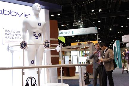 A trade show booth with a large white mannequin on the left side of the booth. The booth has a sign that reads "Abbvie" and "Do your patients have HCV?" with three circular stickers on it. On the right side there are two men standing in front of the display one of them is holding a cup of coffee and the other is looking at the display. In the background there is a large banner with the word "Intercept" and a logo for the company. The overall atmosphere of the image is professional and modern.