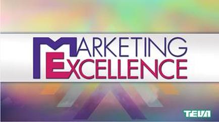A logo for a company called "Marketing Excellence". The logo is rectangular in shape and has a white background. The word "MARKETING EXCELLENCE" is written in bold capital letters in a modern sans-serif font. The letters are arranged in a diagonal line with the "M" in the center and the "E" on the left side. The "X" is slightly larger than the other letters and is slightly smaller than the rest of the text. The background is blurred but it appears to be a gradient of colors with a rainbow-like effect. The logo also has the word "tea" written in a smaller font at the bottom right corner.