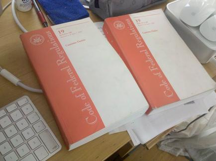 Two books on a desk. The books are red and white in color and have the title "Code of Federal Regulations" written on the cover. The cover also has the number "19" written in red. The desk is cluttered with various items including a keyboard a mouse a pen a blue cloth and a white box. There are also some papers scattered around the desk.