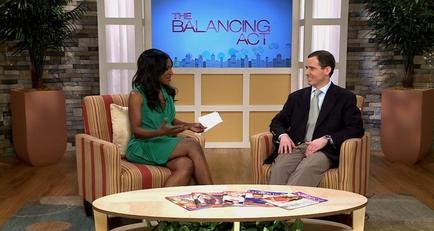 A man and a woman sitting in a TV studio. The woman is wearing a green dress and is holding a piece of paper in her hand. She is sitting on a striped armchair with a coffee table in front of her. The man is sitting next to her and is also wearing a suit and tie. They are both smiling and appear to be engaged in a conversation. In the background there is a large screen with the words "The Balancing Act" and a city skyline. There are two potted plants on either side of the screen.