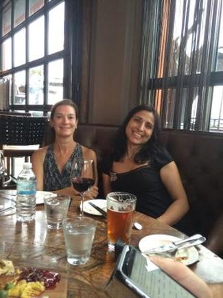 Two young women sitting at a table in a restaurant. They are both smiling and appear to be enjoying their meal. The table is set with plates glasses and a menu card. The woman on the left is wearing a black top and has long dark hair. She is holding a glass of red wine in her hand and is looking at the camera. The background shows a large window with a view of trees and buildings outside.