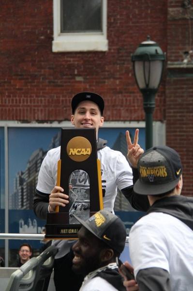 A young man holding up a large trophy with the NCAA logo on it. He is wearing a black baseball cap and a white jacket with the word "NCAA" written on it in gold letters. He has a big smile on his face and is making a peace sign with his right hand. There are two other men in the background one of whom is also holding up the trophy. The background shows a brick building and a street lamp. It appears that the young man is celebrating a victory or victory.