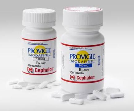 Two white plastic bottles with red and blue labels. The labels have the brand name "prodigal Modafinil" written in bold black letters at the top followed by the product name "100 mg" in smaller black letters. Below that there is a label that reads "100 Tablets" in red letters. The bottle on the left is labeled "cephalic" and has a red cap. On the right is labeled with "200 mg" and "100 tablets". There are also a few white pills scattered around the bottles. The background is plain white.