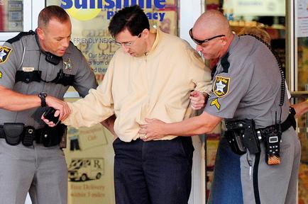 Three police officers in uniform standing in front of a store window. The man in the center is wearing a beige shirt and glasses and appears to be in his late twenties or early thirties. He is being escorted by two of the officers on either side of him. The officer on the left is holding the man's hand while the man on the right is holding his arm around his waist. All three officers are wearing their uniforms with badges and bulletproof vests. The store window behind them has a sign that reads "Sunshine Pharmacy" and there is a picture of a car on it.