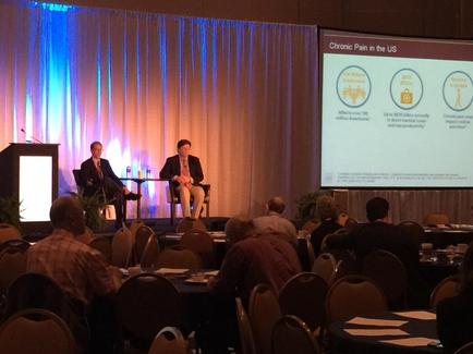 Two men in business suits are sitting on a stage facing an audience. A lectern is to the left of the image and a presentation screen (with a slide on Chronic pain in the U.S.) is to the right of the image.