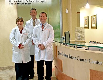 Three people two men and a woman standing in front of a reception desk at the James Graham Brown Cancer Center. They are all wearing white lab coats and are smiling at the camera. The desk has a sign that reads "Dr. Lynn Parker Dr. Daniel Metzinger and Dr. Michael milan" and there are several framed pictures hanging on the wall behind them. The room appears to be well-lit with natural light coming in from the windows.