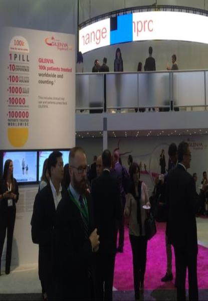A group of people gathered in a large room with a pink carpet. The room appears to be an exhibition or trade show booth as there is a large banner on the left side of the image that reads "100% PILL" and a sign on the right side that says "glenda 100% patients treated worldwide and counting". There are several people in the background some of whom are standing and some are walking around the booth. The people are dressed in formal business attire and appear to be engaged in conversation.