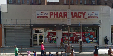 A large pharmacy building with a red and white sign that reads "Sugar Hill Pharmacy". The building is located on a street corner with a sidewalk in front of it. There are several people walking on the sidewalk some of them carrying shopping bags while others are sitting on benches. The pharmacy has a large window display with various items for sale including a red truck and a white van. The building appears to be old and dilapidated with peeling paint and broken windows. The sky is overcast and there are other buildings visible in the background.