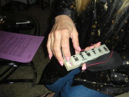 A person's hand holding a small white box with the letters "MTWTFS" written on it. The person is wearing a black jacket with gold stars on it and blue jeans. They are sitting on a chair with a purple piece of paper and a remote control in front of them. The background appears to be a living room or a bedroom.
