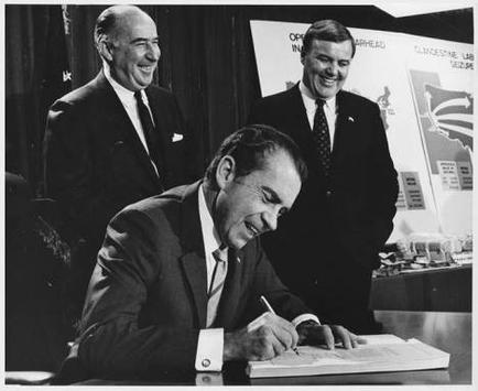 A black and white photograph of three men in formal attire. The man in the center is sitting at a desk and is signing a document with a pen. He is wearing a suit and tie and appears to be in his late 60s or early 70s. The other two men are standing behind him also wearing suits and ties. They are all smiling and looking at the man signing the document. In the background there is a banner with the logo of the United States Air Force and a map of the world.