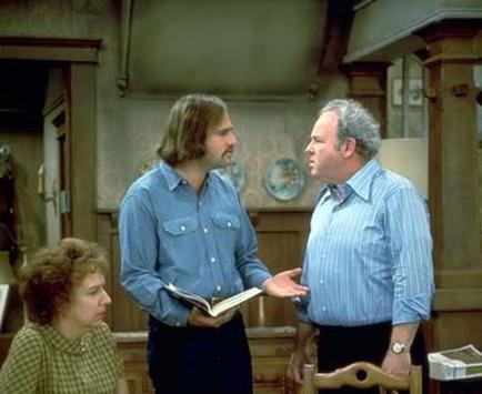 Three people in a kitchen. The person on the left is a man with long hair and a beard wearing a blue button-down shirt. He is holding a book in his hand and appears to be explaining something to the person in the middle who is a woman with curly hair. The man on the right is an older man with grey hair and is wearing a light blue striped shirt. They are standing in front of a wooden cabinet and there is a window in the background. The woman is sitting on a chair and is looking at the man with a concerned expression on her face.