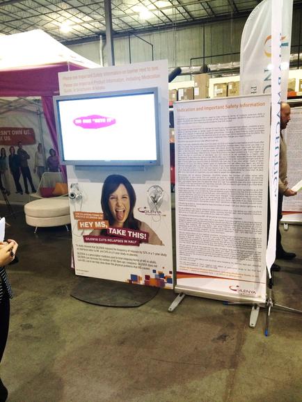 A large white banner stand with a large screen on top of it. The screen displays an image of a woman's face with the text "Hey Ms. Take This!" above her head. Below the screen there is a text that reads "Welcome to our safety information". On the right side of the banner stand there are two smaller banners with text that read "Safety Information". The background of the image shows an indoor space with other people and tents.
