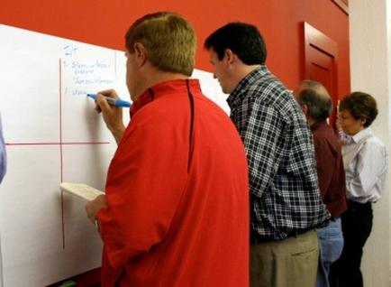 A group of people standing in a room with red walls. In the center of the image there is a man wearing a red jacket and holding a blue marker and writing on a whiteboard. He appears to be giving a presentation or brainstorming session. To his right there are two other men one wearing a black and white checkered shirt and the other wearing a blue shirt. They are both looking at the whiteboard and appear to be engaged in the conversation. There is a door visible in the background.