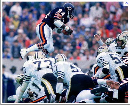 A football game in progress between the Chicago Bears and the New Orleans Saints. The Bears player number 34 is in the air holding the football and running towards the end zone. He is wearing a blue and orange jersey with the number 34 on it and a black helmet. The Saints players are wearing white and gold uniforms with the numbers 73 and 75 on them. The background shows a crowd of spectators in the stands. The image appears to have been taken during a game as the players are in the midst of a tackle attempt.