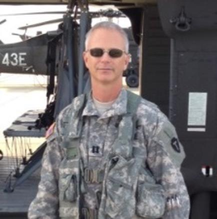 A man in a military uniform standing in front of a helicopter. The man is wearing a camouflage uniform with a badge on his left chest and a backpack on his right shoulder. He is also wearing sunglasses and has a serious expression on his face. The helicopter in the background has the number 43E written on it. The sky is blue and the ground appears to be an airport tarmac.