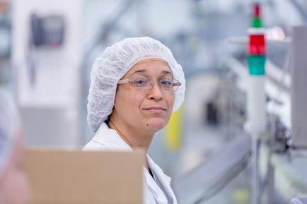 A woman wearing a white lab coat and a white hairnet on her head. She is standing in a laboratory or laboratory setting with various machines and equipment in the background. The woman is looking directly at the camera with a serious expression on her face. She appears to be a scientist or a researcher as she is wearing glasses and has a clipboard in front of her. The background is blurred but it seems to be an industrial setting with bright lights and machinery.