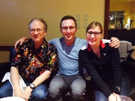Three people sitting on a couch in a living room. On the left is an older man wearing a colorful Hawaiian shirt and glasses. He is sitting next to a younger man in a blue shirt and gray pants. The younger man is in the middle with his arms around the older man's shoulders. All three people are smiling and appear to be happy. The woman on the right is wearing a black jacket and glasses and she is also smiling. In the background there is a coffee table with a glass of wine and a plate of food.
