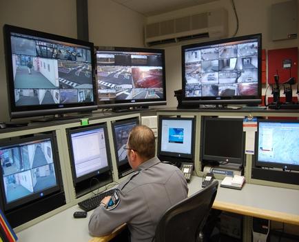 A man sitting at a desk in front of multiple computer monitors. The monitors are arranged in a row and each monitor displays a different image or video. The man is wearing a gray uniform and appears to be a security guard or a police officer. He is sitting at the desk with his back to the camera and is looking at the monitors intently. The desk is cluttered with various items such as a keyboard mouse telephone and other office supplies. The background of the image shows an air conditioning unit and a red bulletin board.