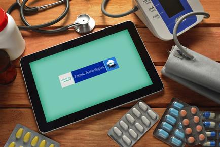 A tablet with a green screen displaying the logo of Patient Technologies. The tablet is placed on a wooden table with various medical supplies scattered around it. On the right side of the image there is a blood pressure monitor a stethoscope and a bottle of medicine. Next to the tablet there are several blister packs of pills. The background is blurred but it appears to be a medical setting.