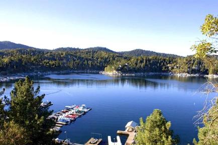 A beautiful landscape of a lake surrounded by trees and mountains. The lake is calm and the water is a deep blue color. On the right side of the image there is a wooden dock with several boats docked at it. The boats are of different sizes and colors including red white and blue. In the background there are more trees and hills covered in green foliage. The sky is clear and blue and the overall scene is peaceful and serene.