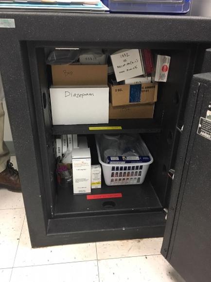 A black metal cabinet with a door open. Inside the cabinet there are several items neatly arranged. On the top shelf there is a white box with the word "Diazepam" written on it. Next to the box it appears to be a stack of cardboard boxes with labels on them. In the bottom shelf it seems like there are a few items in a plastic basket. The cabinet is located in a room with a tiled floor and a person's leg visible in the background.