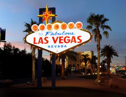 A photograph of the iconic "Welcome to Fabulous Las Vegas Nevada" sign. The sign is a large red and white sign with a star in the center and the words "Welcome" written in bold capital letters. The word "Fabulous" is written in a larger font size than the rest of the text. The background of the sign is lit up with colorful lights creating a vibrant and lively atmosphere. The sign is located on a street lined with palm trees and other buildings and there are a few cars parked on the side of the road. The sky is a deep blue and the sun is setting in the distance casting a warm glow over the scene. The overall mood of the image is cheerful and welcoming.