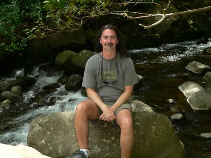 A man sitting on a large rock in front of a stream. He is wearing a gray t-shirt shorts and sneakers. He has long dark hair and a beard and is smiling at the camera. The stream is flowing over rocks and there are trees and bushes in the background. The water is crystal clear and the rocks are jagged and uneven. The man appears to be relaxed and enjoying the view.