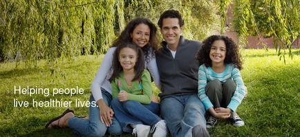 A family of four sitting on a grassy lawn in a park. The family consists of a man a woman and two young girls. The man is in the center with the woman on his left and the two girls on his right. They are all smiling and looking at the camera. The woman is wearing a white top and blue jeans while the girls are wearing colorful tops and jeans. The background shows trees and a brick building. On the left side of the image there is text that reads "Helping people live healthier lives."