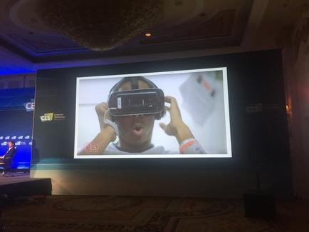 A large screen in a conference room with a chandelier hanging from the ceiling. On the screen there is an image of a person wearing a virtual reality headset. The person is holding the headset up to their eyes and appears to be in the middle of a video call. The background of the screen is white and there is a blue banner on the left side of the image with the CES logo. The room has a carpeted floor and a stage on the right side.