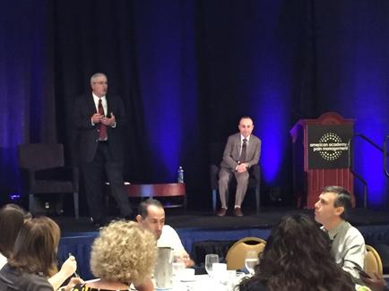 A stage with a blue curtain and a podium on the right side. On the left side of the stage there is a man standing and speaking into a microphone. He is wearing a suit and tie and appears to be giving a presentation. In front of him there are two men sitting on chairs facing each other. The man on the left is holding a microphone and is gesturing with his hands as he speaks. The audience is seated at a table with white tablecloths and is attentively listening to the presentation. There are several people sitting at the table some of whom are looking at the man speaking.