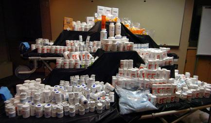 A large number of white plastic containers stacked on top of each other on a black table. The containers are arranged in a pyramid-like shape and appear to be of different sizes and shapes. Some of the containers are filled with orange and white containers while others are empty. There are also some plastic bags and boxes scattered around the table. In the background there is a whiteboard and a projector screen suggesting that the image was taken in a room with a projector and other equipment.