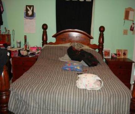 A bedroom with a wooden bed in the center. The bed has a wooden headboard and footboard with intricate carvings. The walls are painted in a light green color and there is a black curtain hanging on the wall above the bed. On the left side of the bed there are two wooden nightstands with various items on them including a dresser with a lamp a picture frame and a few other decorative items. On top of the dresser there is an open book and a stuffed animal. The floor is covered with a gray and white striped comforter. The room appears to be messy and cluttered.
